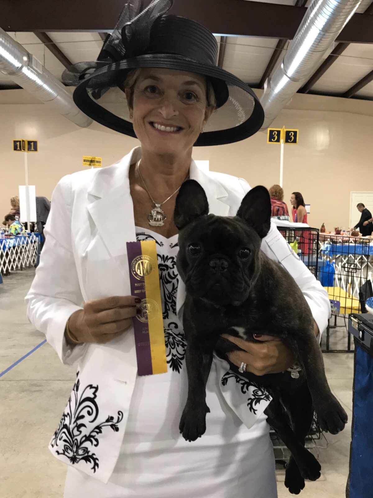 Woman in black hat and white suit holds black French bulldog and award ribbons.