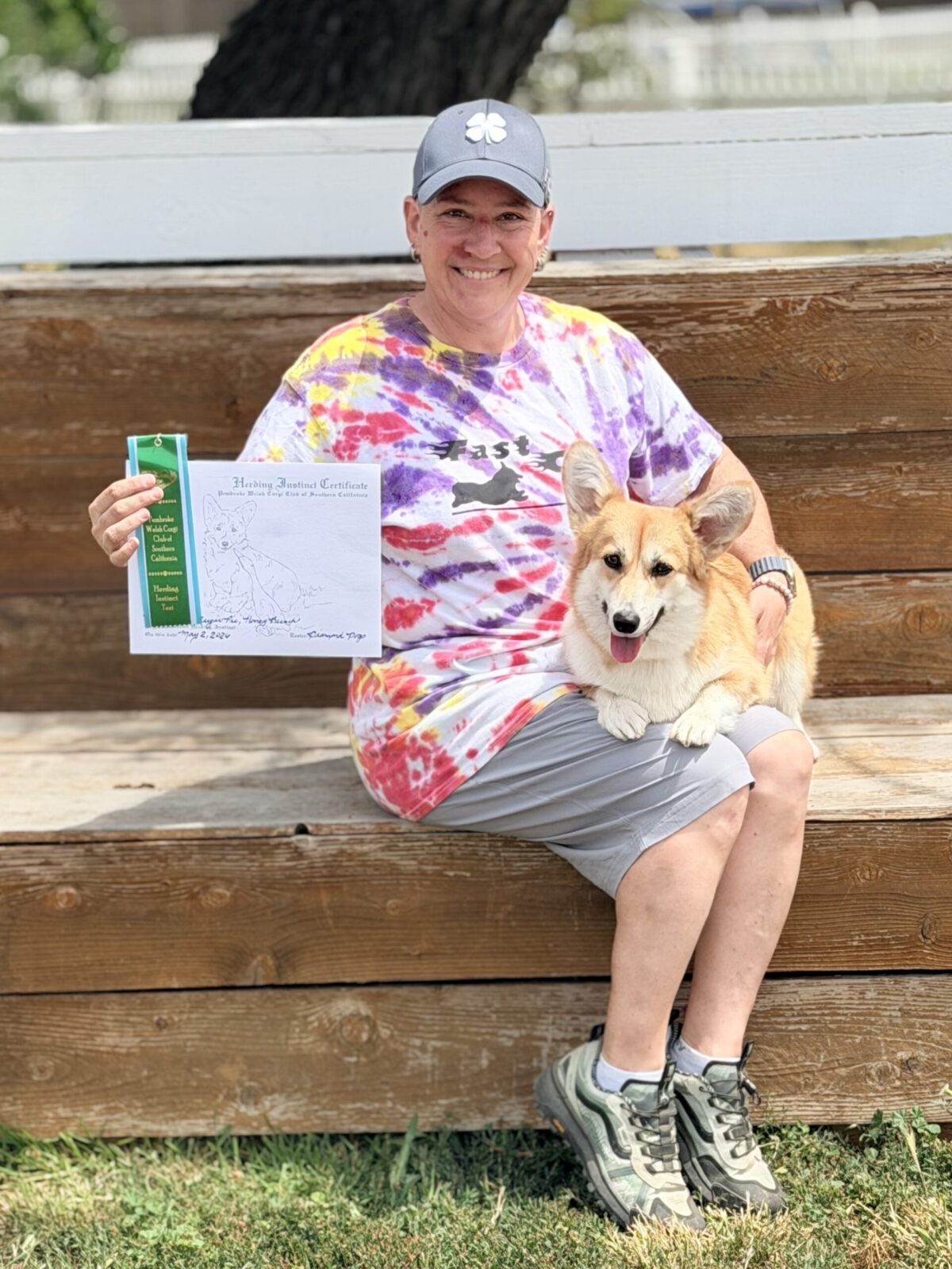 Woman in tie-dye shirt and cap holds a certificate and a corgi dog.