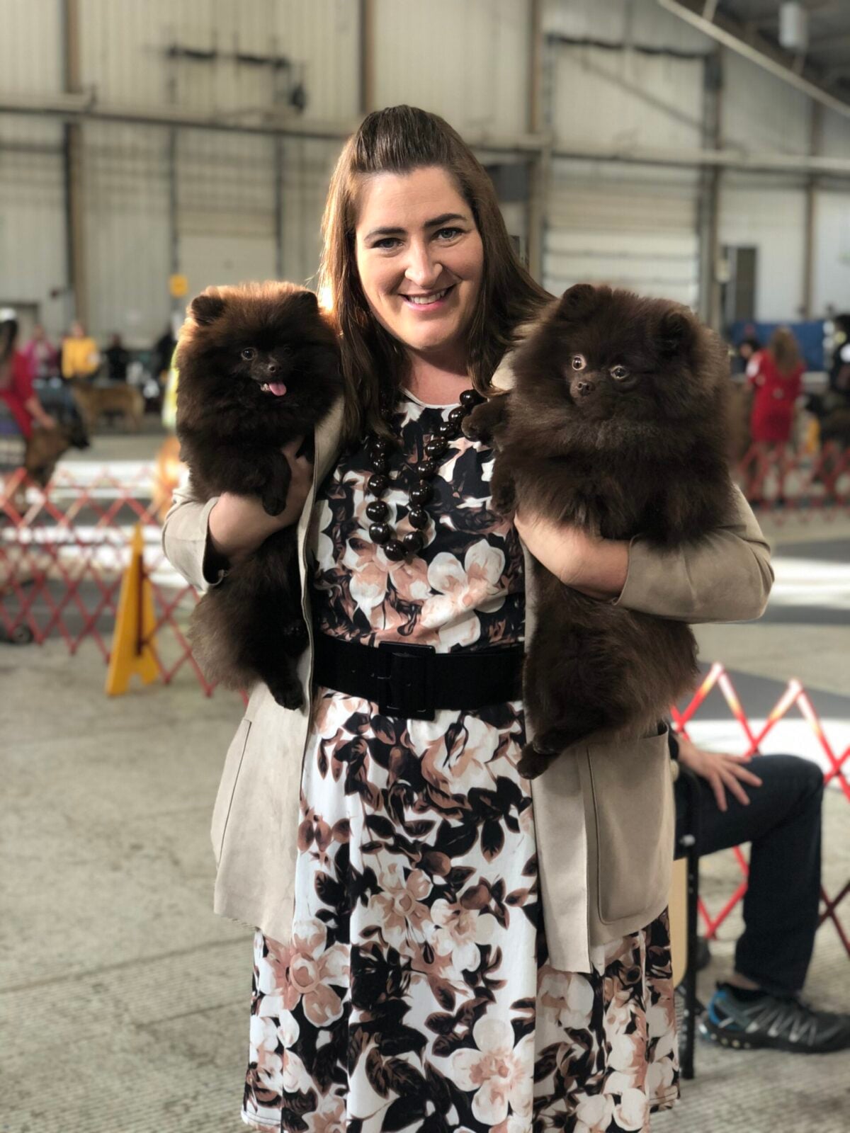 Woman holding two fluffy brown Pomeranian puppies at a dog show.