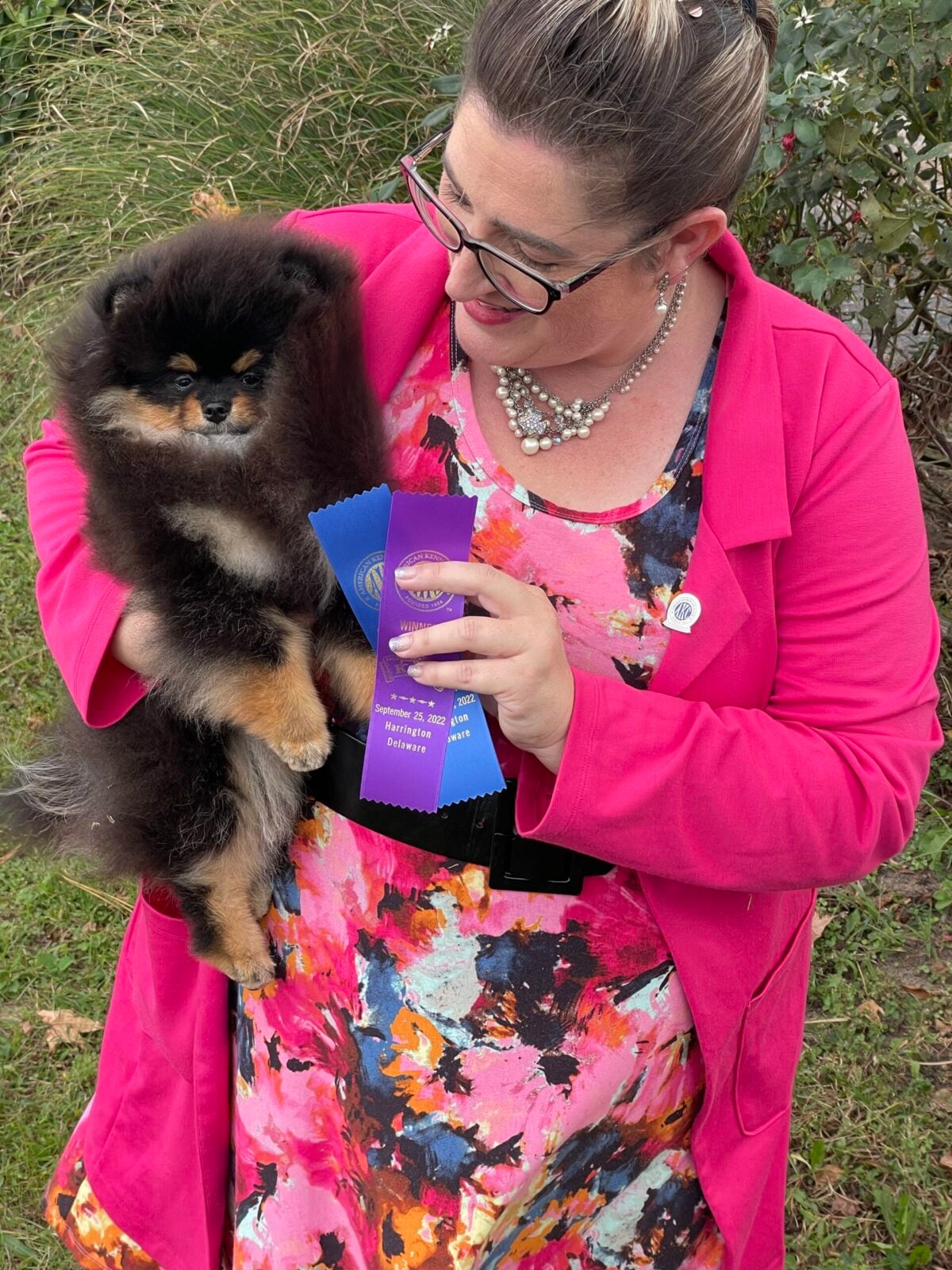 Woman in pink jacket holding fluffy black Pomeranian puppy with blue and purple ribbons.