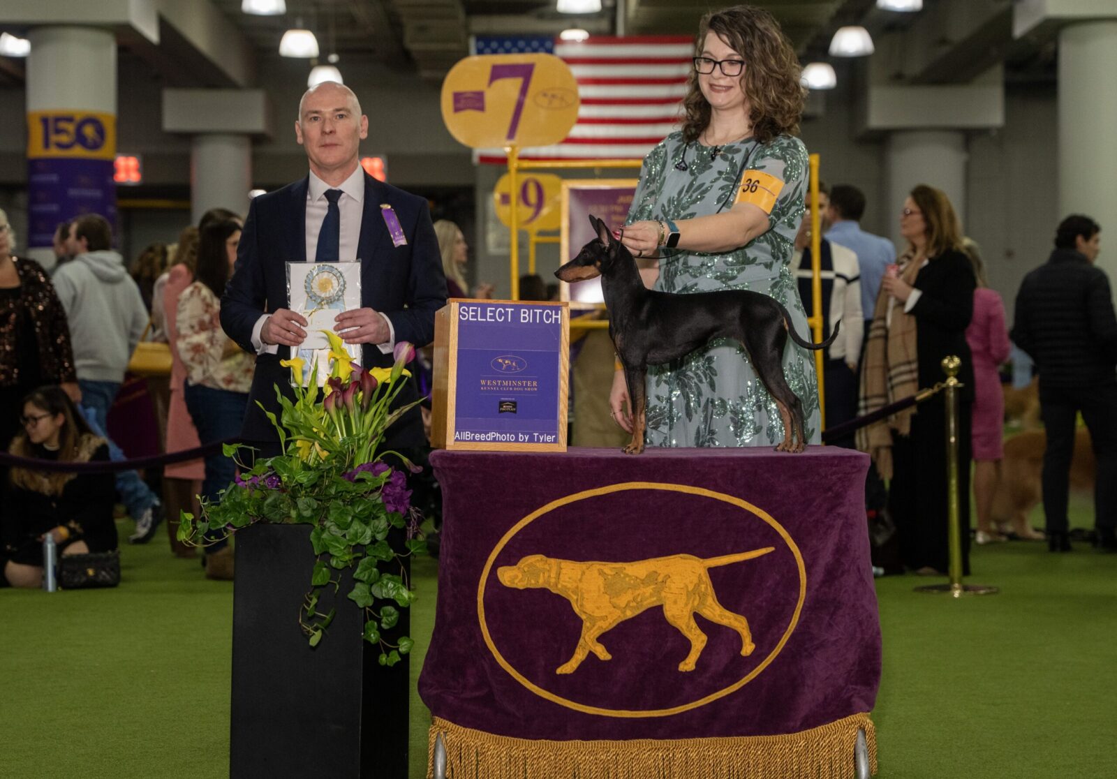 A man holds an award beside a woman with a black dog on a podium, surrounded by a colorful audience at a dog show.