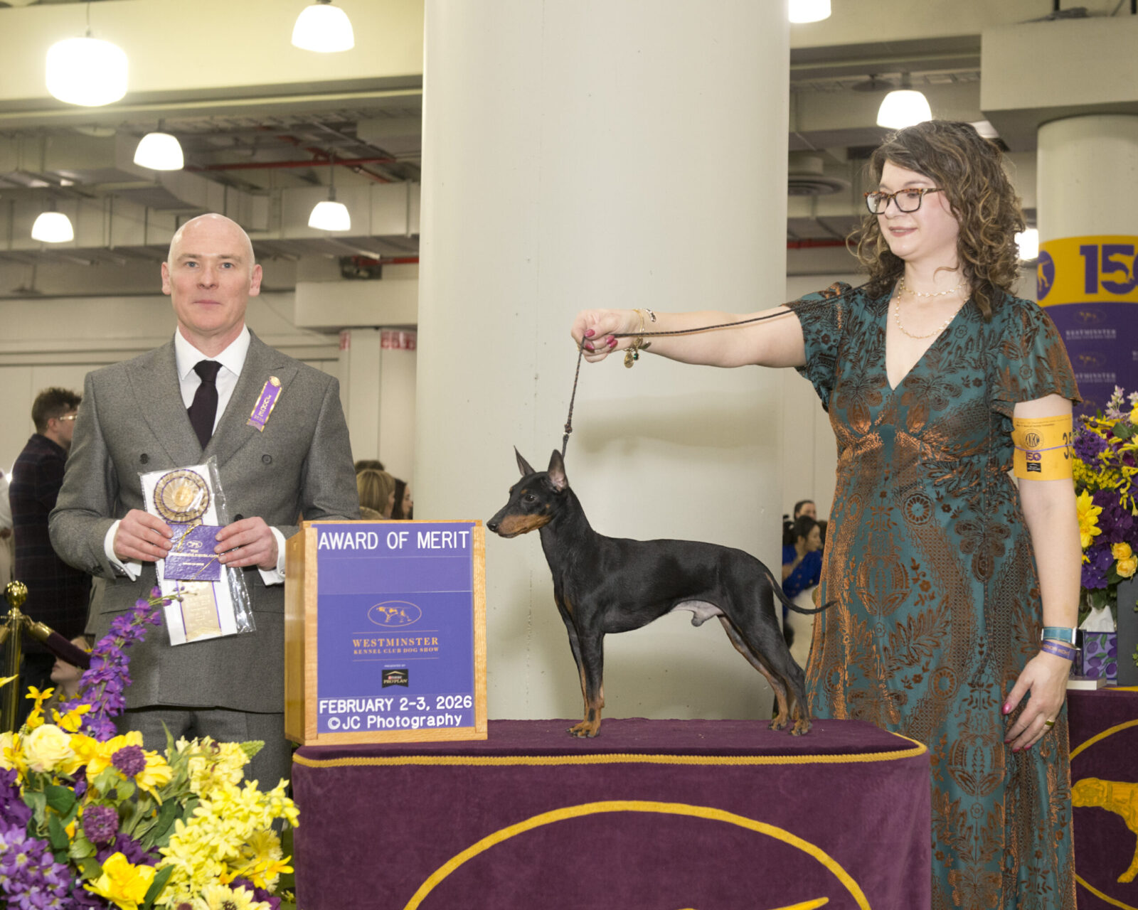 A woman in a green dress holds a leash attached to a small dog, while a man in a suit displays an award at a dog show.