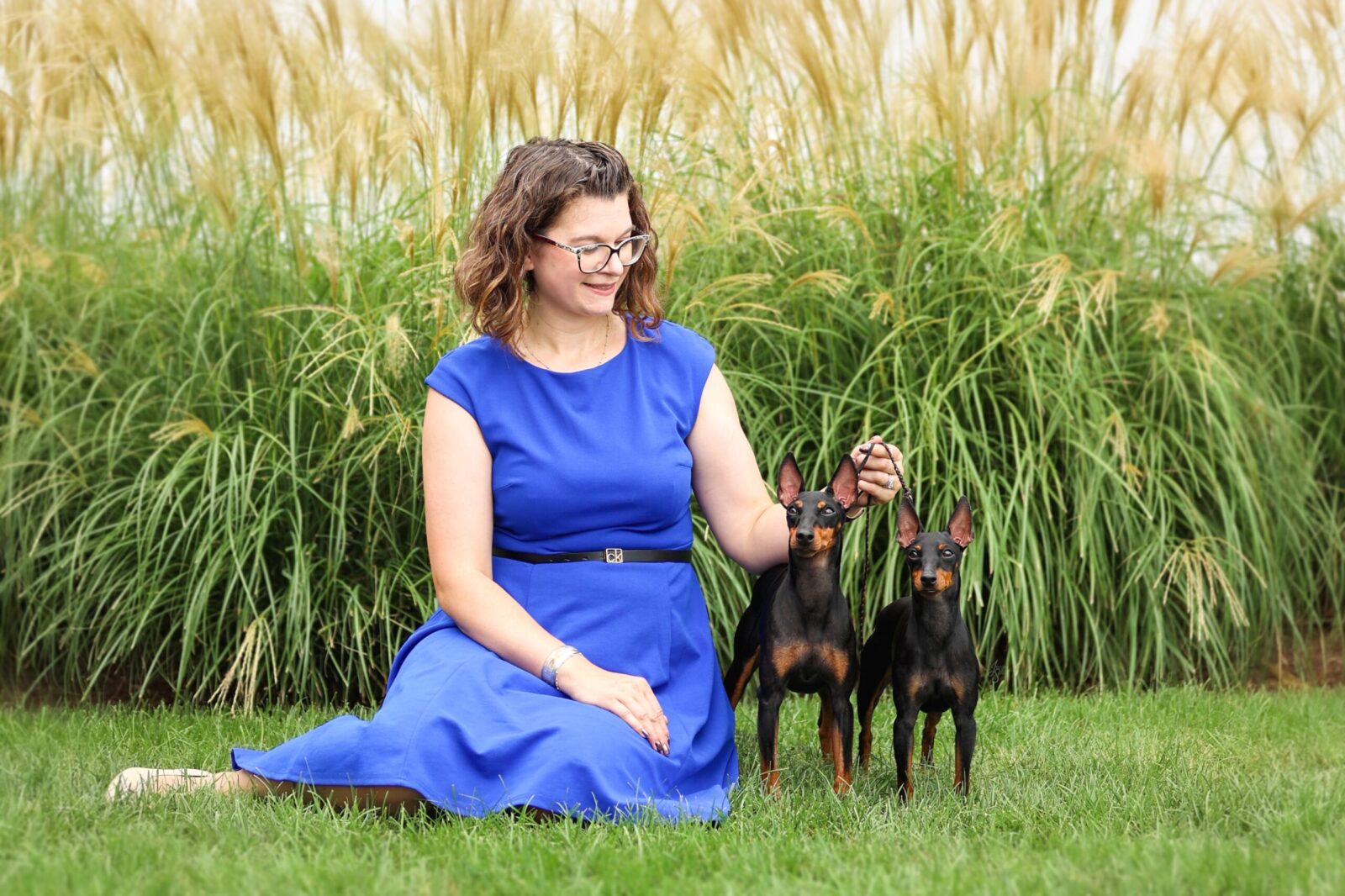 A woman in a blue dress sits on grass, smiling with two black and tan dogs beside her, framed by tall green grass.