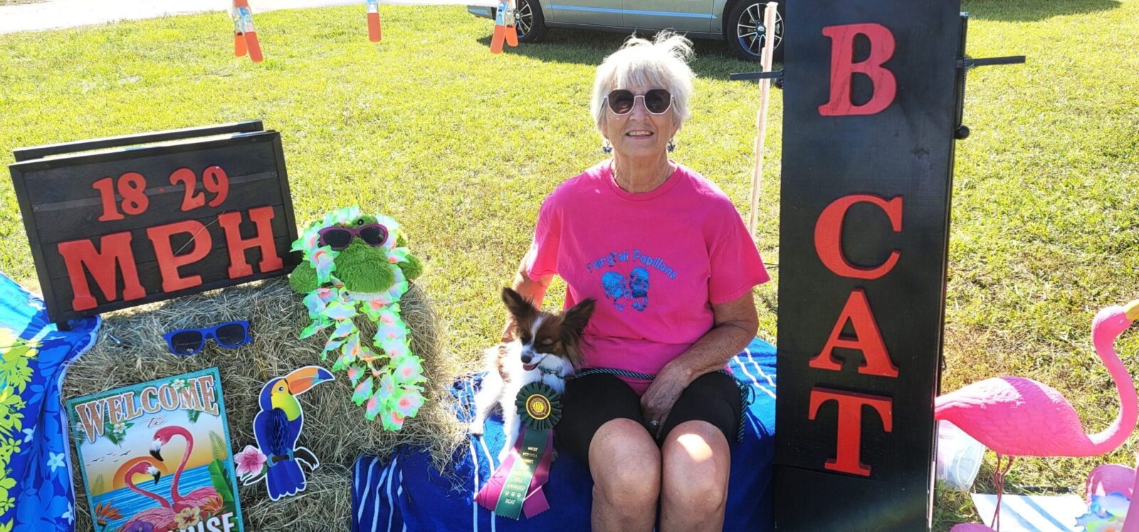 A smiling woman in a pink shirt sits with a small dog, surrounded by colorful decorations and a sign reading "18-29 MPH."