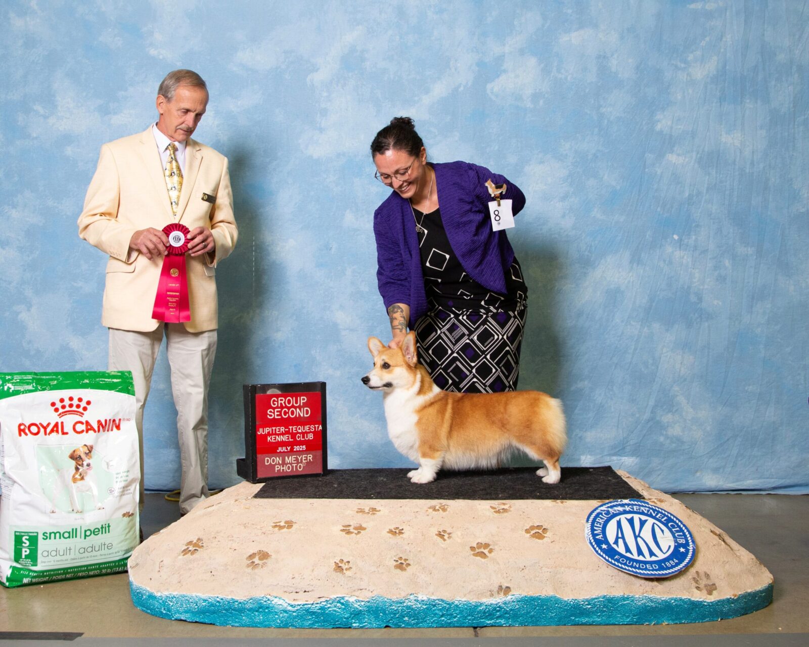 Corgi on a podium with a handler, a judge holding a red ribbon, and a sign displaying "Group Second."