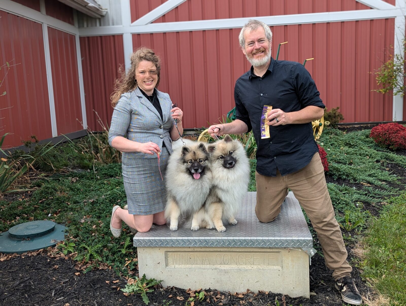 A woman in a gray dress and a man in a black shirt pose with two fluffy dogs on a concrete platform in a garden setting.