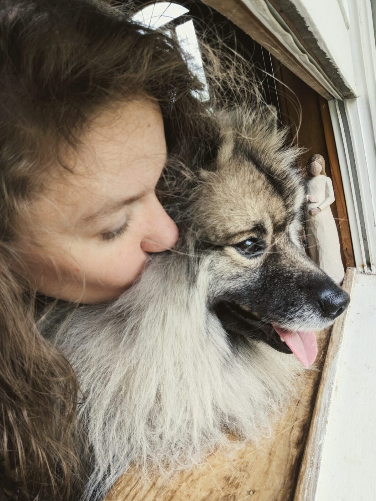 A woman leans in to kiss her fluffy dog, both resting their heads against a window with soft natural light.