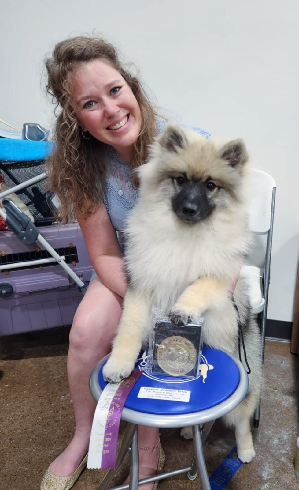 A smiling woman holds a fluffy dog beside a medal and ribbons on a blue stool in a bright indoor setting.