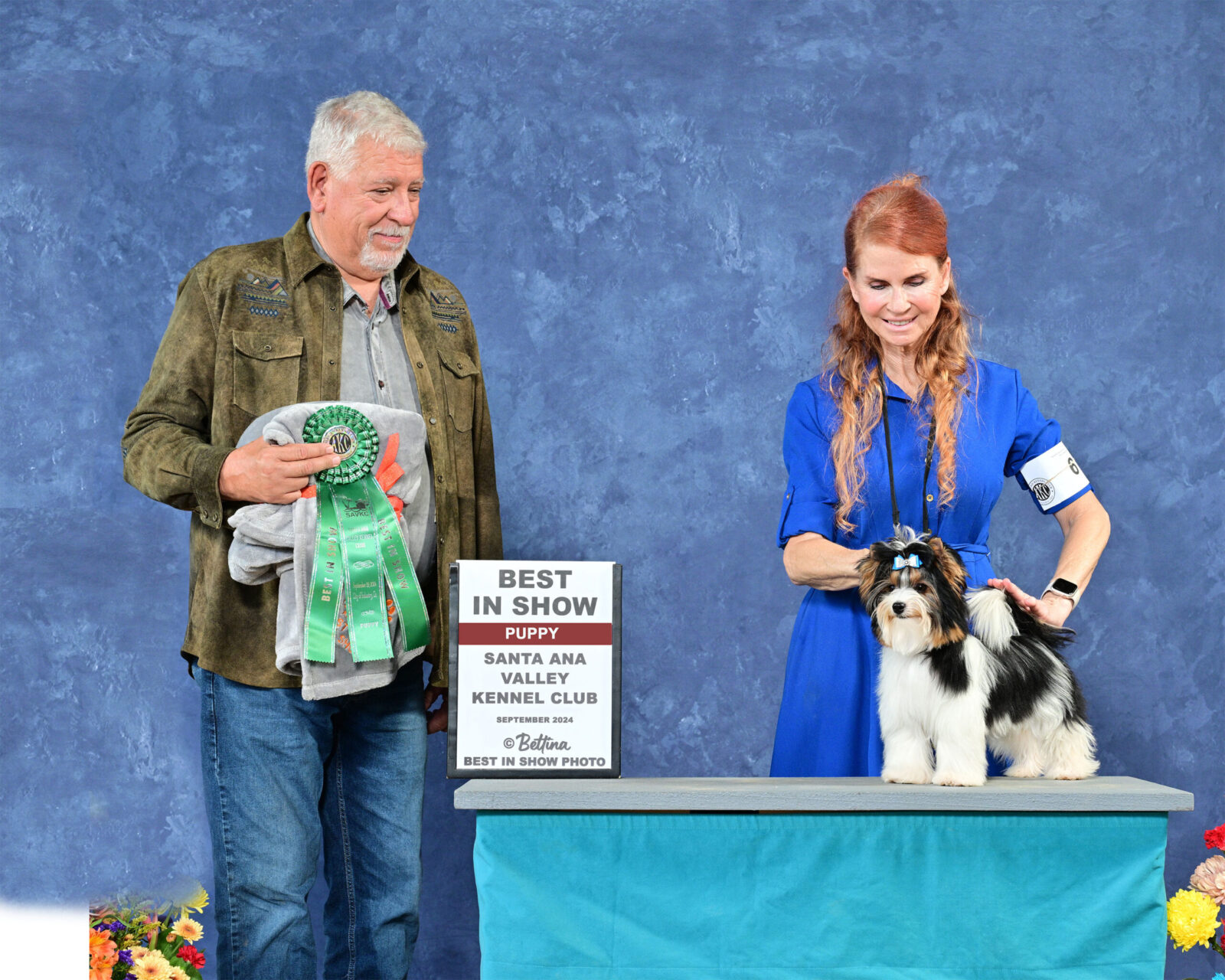 A man holds a trophy while a woman presents a small dog on a table, both smiling against a blue backdrop.