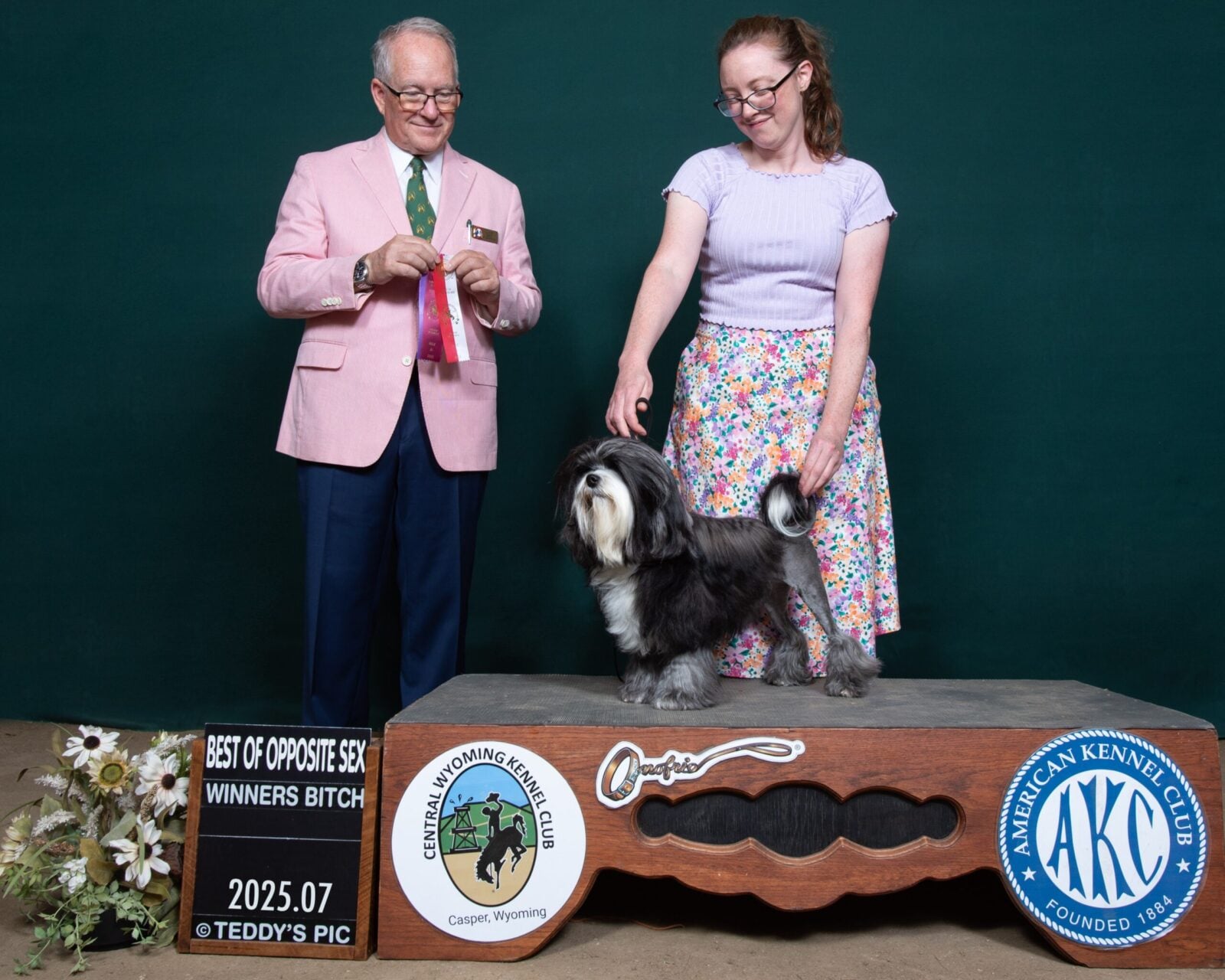 A woman in a floral skirt presents a small dog on a podium, while a man in a pink blazer holds a ribbon.