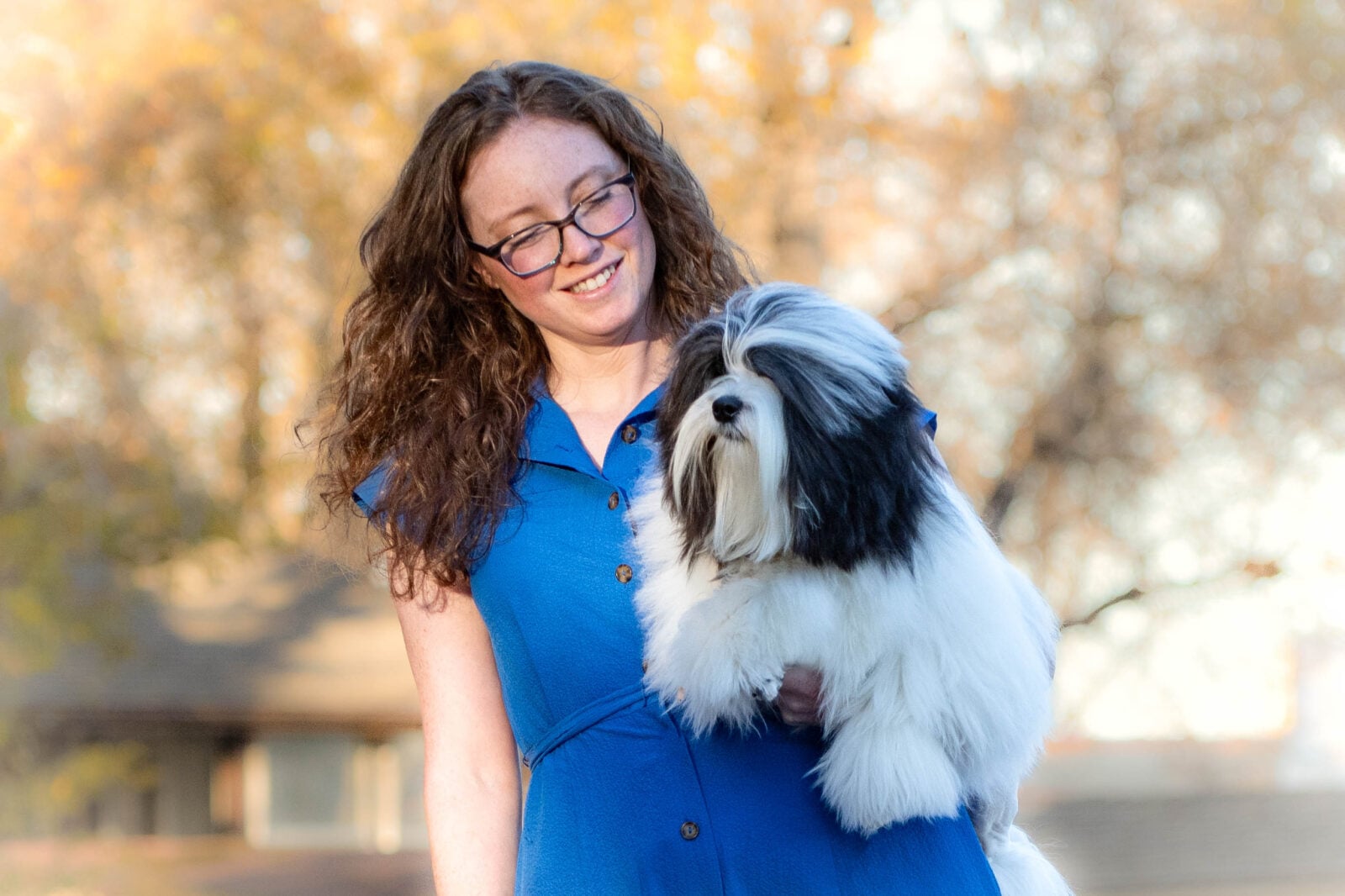 A woman in a blue dress smiles while holding a fluffy black-and-white dog against a blurred autumn background.