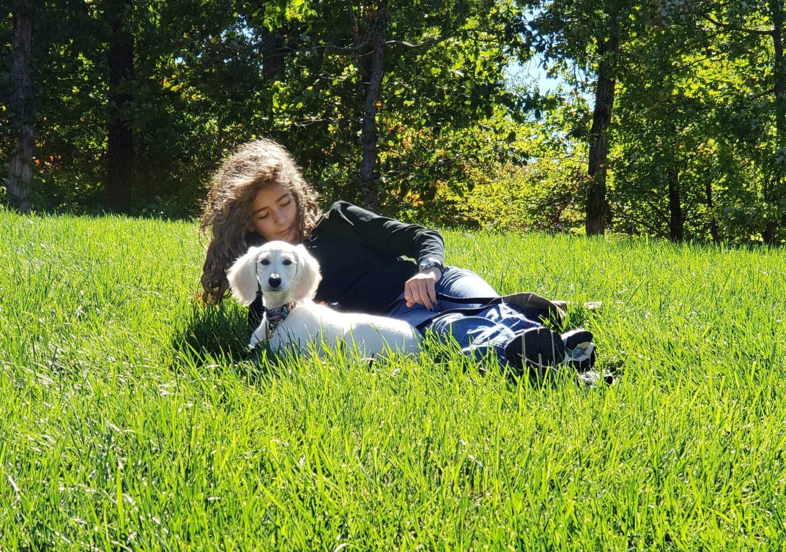 Young person and white dog lying in lush green grass under trees.