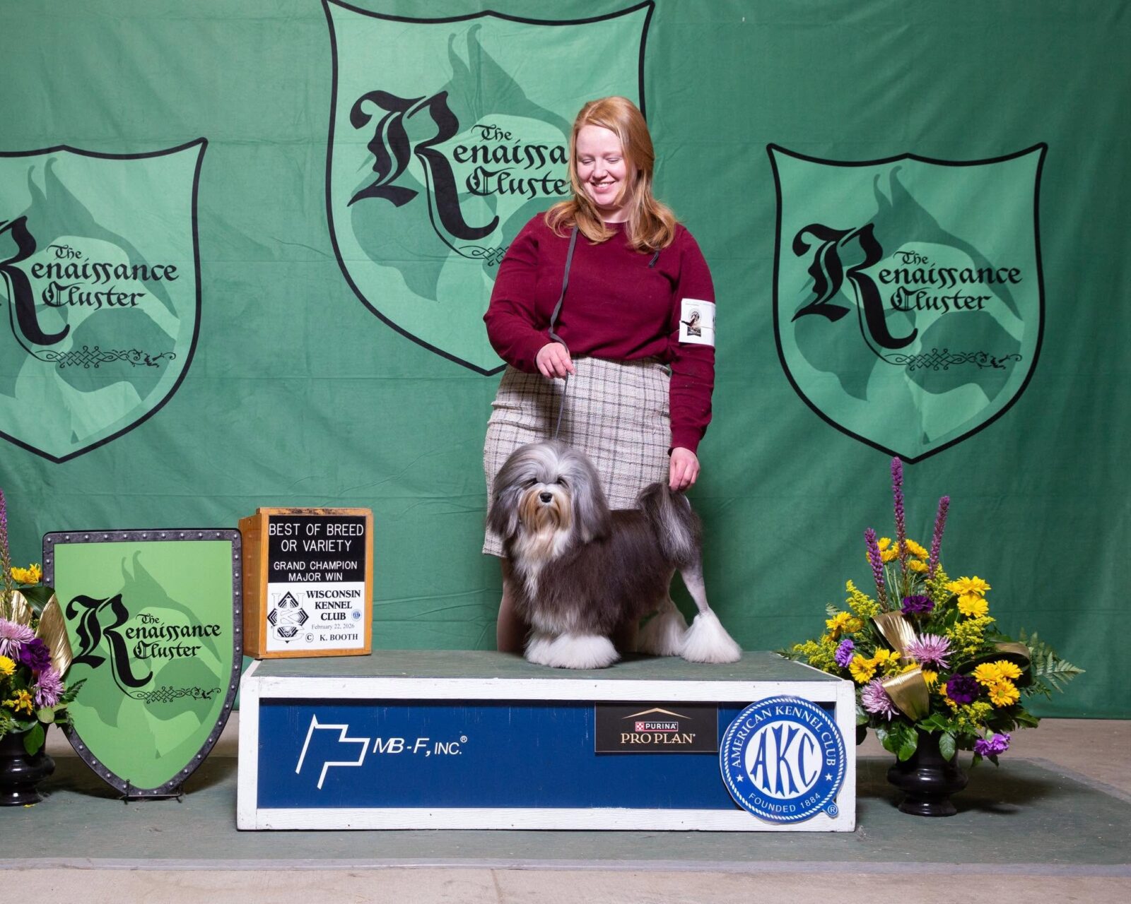 Woman with a Havanese dog on a podium, with awards and floral arrangements.