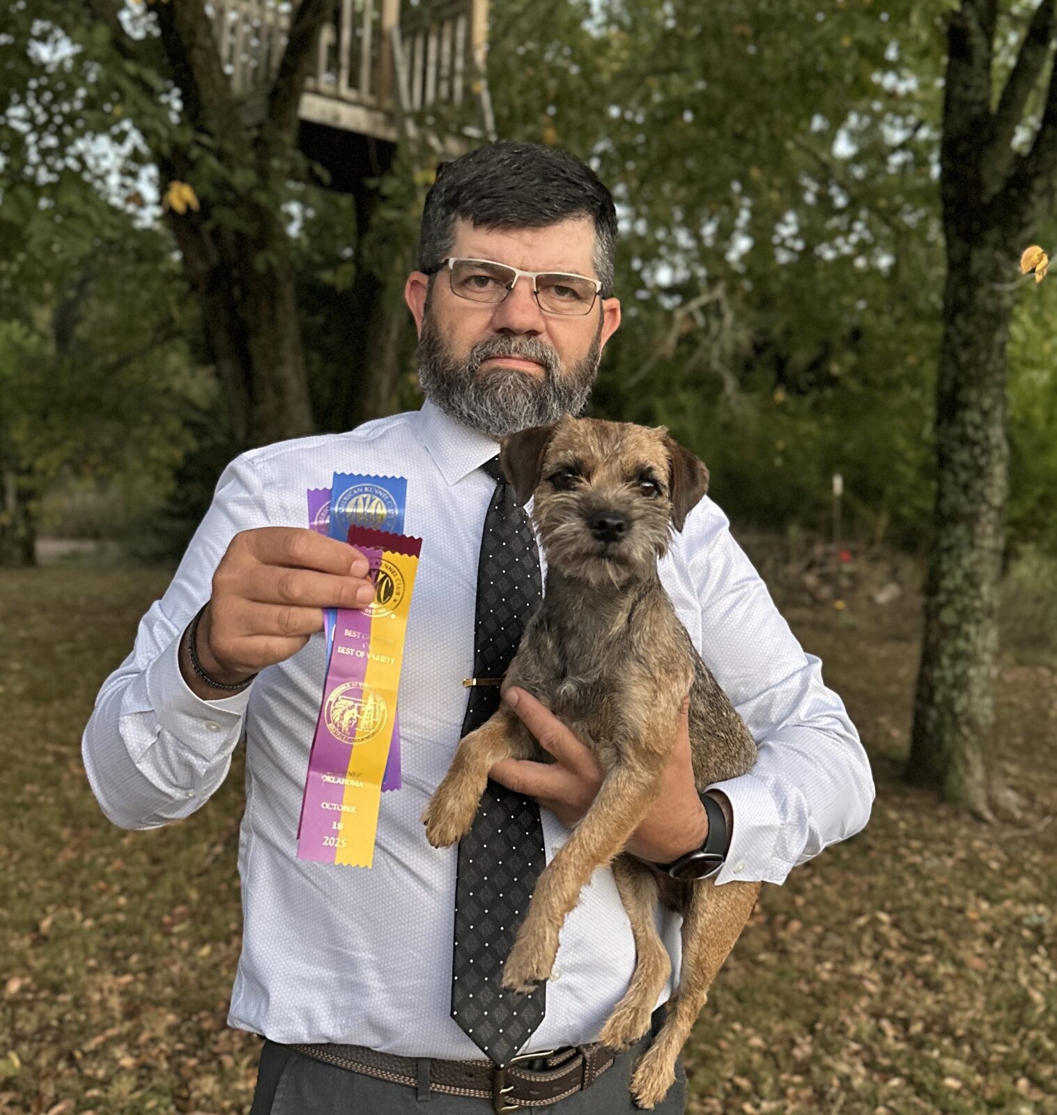 Man holding a Border Terrier and three award ribbons outdoors.